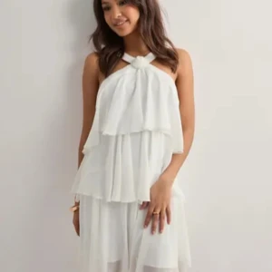 Vit volangklänning med öppen rygg - Superfin vit kortklänning med flera lager volanger och snygg halterneck med rosettdetalj framtill. Klänningen har öppen rygg och breda axelband, vilket ger en luftig och somrig känsla. Perfekt för dig som vill sticka ut med en romantisk och trendig look.Nypris: 69.99eur.