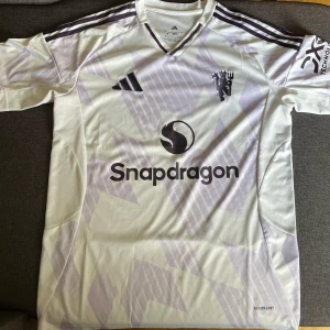 Manchester United bortatröja Adidas - Snygg Manchester United bortatröja från Adidas med Snapdragon sponsortryck. Tröjan är vit med ljuslila geometriskt mönster, svarta detaljer och klubbens emblem på bröstet. Tillverkad i lätt och ventilerande polyester med korta ärmar och klassiska Adidas-ränder på axlarna. Storleken är S 