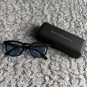  Corlin Eyewear Solglasögon  - Snygga svarta glasögon från Corlin Eyewear med klassisk rundad form och blåtonade glas. Kommer med originalfodral. Perfekta för dig som vill ha en stilren och modern look.