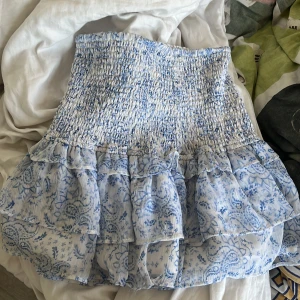 Blåvit volangkjol från Gina Tricot - Supersöt kort kjol från Gina Tricot i blått och vitt med smockad midja och flera volanger. Kjolen har ett blommigt mönster och är gjord i ett lätt, luftigt material som passar perfekt till varma dagar. Perfekt för dig som gillar en romantisk och somrig stil!
