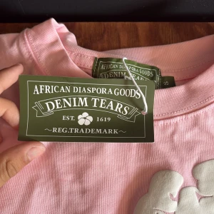 Rosa blommig t-shirt från Denim Tears - Säljer en rosa t-shirt från Denim Tears med stora vita blommor i 3D-tryck över hela tröjan. T-shirten har klassisk passform, rund hals och korta ärmar. Perfekt statement-plagg för dig som vill sticka ut.
