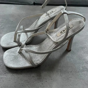 Silvriga klack-sandaler från NA-KD - Snygga silvriga sandaler från NA-KD med smala remmar och hög klack. Storlek 39. Skorna har en texturerad yta som ger en lyxig känsla och en fyrkantig tå. Perfekta för dig som vill sticka ut med stilrena detaljer.