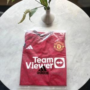 Manchester United Adidas matchtröja S - Manchester Uniteds officiella matchtröja från Adidas i rött med svarta detaljer och klubbmärke på bröstet. Tröjan har korta ärmar, vit TeamViewer-logga och diskret geometriskt mönster i tyget. Tillverkad i lätt och ventilerande material (AEROREADY).