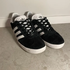Adidas Gazelle svarta sneakers mocka - Klassiska Adidas Gazelle sneakers i svart mocka med vita detaljer och de ikoniska tre ränderna på sidorna. Skorna har vit sula, svart snörning och vit plös med Adidas-logga. Perfekta för dig som gillar retrostil och streetwear. Använda fåtal gånger men mycket fina i skicket.