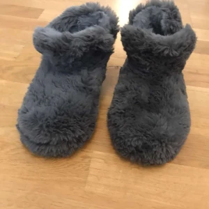 Grå fluffiga tofflor storlek 37/38 - Supermysiga grå tofflor med fluffig pälsimitation och mjuk insida. Perfekta att glida runt i hemma när det är kallt. Sulan är i syntetmaterial och har greppmönster undertill. Tofflorna är höga och täcker ankeln för extra värme.