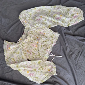 Blommig croppad blus från Aware - Supersöt croppad blus från Aware med smockad kropp och långa puffärmar. Toppens vita tyg har ett blommigt mönster i pastelliga toner av rosa, lila, gul och grönt. Knytband i midjan och volangdetaljer vid halsen ger en romantisk vibe. Perfekt för vår och sommar!