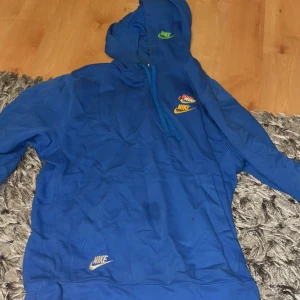 Blå Nike hoodie med broderade loggor - Säljer en blå hoodie från Nike med flera broderade loggor i olika färger på bröstet, huvan och ryggen. Tröjan har huva med snörning, känguruficka och långa ärmar. Perfekt för dig som gillar streetwear och vill sticka ut lite extra.