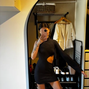 Svart cut out klänning från NA-KD - Säljer en svart kort klänning från NA-KD med asymmetrisk design, en lång ärm och en ärmlös sida. Klänningen har hög hals och en snygg cut out-detalj vid midjan. Tight passform och stretchigt material som sitter snyggt på kroppen.