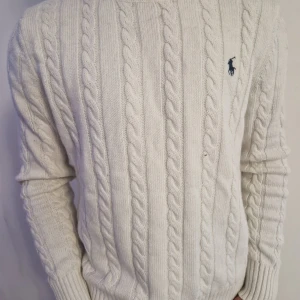 Polo ralph lauren sweater - Kabelstickad vit tröja från Polo Ralph Lauren med klassisk rund hals och broderad marinblå logga på bröstet. Tröjan är långärmad och har ribbade muddar och nederkant. Perfekt för dig som gillar stilren och tidlös design.