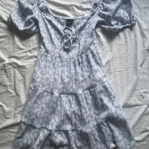 Blommig kortklänning  - Supersöt ljusblå kortklänning från Gina Tricot med vita blommor, puffärmar och snörning framtill. Klänningen har volangdetaljer och en smickrande v-ringning. Perfekt för sommarens alla tillfällen! Man kan välja hur stor v-rignigen ska vara genom att dra hårdare i snörerna💕