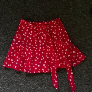 Röd blommig omlottkjol - Superfin röd omlottkjol med vita blommor och volangkant. Kjolen har knytband i midjan och är perfekt för sommaren. Lätt och luftig modell som ger en söt och trendig look. Har innershorts. Aldrig använt!