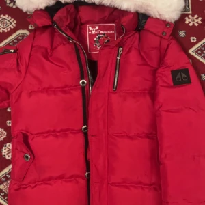 Röd Moose Knuckles dunjacka - Säljer en röd dunjacka från Moose Knuckles med fluffig vit fuskpäls på huvan. Jackan har dragkedja, knappar och flera fickor med snygga detaljer. Perfekt för kalla dagar och har en schysst streetstil med normal passform. Passar bra på 165-175+