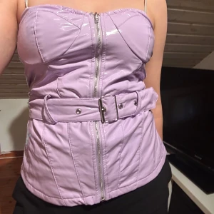 Lila korsett med bälte PrettyLittleThing - Säljer en lila korsett från PrettyLittleThing med glansig finish och markerade sömmar. Toppen har dragkedja framtill och ett brett bälte i midjan med silverspänne. Perfekt för dig som vill sticka ut med en edgy look. Köpt på Sellpy men jag har aldrig använt den💜