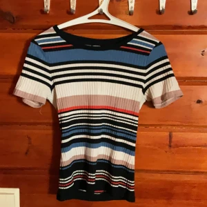 Randig ribbad topp från C XS - Säljer en ribbad topp från C i storlek XS med korta ärmar. Toppen har ett färgglatt randigt mönster i svart, blått, vitt, rött och beige. Den är figurnära och har en rund halsringning. Perfekt för dig som gillar färg och mönster!