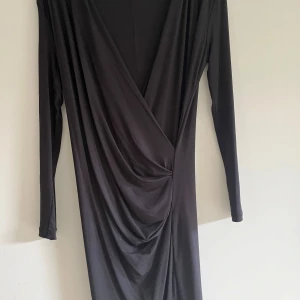 Svart omlottklänning från DAY - Snygg svart långklänning från DAY med omlott-skärning och draperad effekt framtill. Klänningen har lång ärm och djup v-ringning, vilket ger en elegant och stilren look. Perfekt för dig som gillar minimalistisk och trendig design.