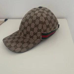 ÄKTA Gucci keps med GG-mönster, storlek L - Snygg keps från Gucci i beige och brunt med klassiskt GG-monogram över hela kepsen. På sidan finns den ikoniska gröna och röda randen. Justerbar rem baktill i mörkbrunt skinn. Perfekt accessoar för att lyfta din outfit. Några  trådar på baklädret sticker ut och kepsen är lite solbränd, men nypris ligger på runt 4500kr