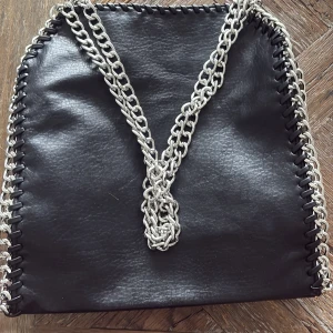 Svart axelväska med silverkedja - Snygg svart axelväska med grova silverfärgade kedjor som axelrem och dekor längs kanterna. Väskan har en modern, kantig form och är tillverkad i skinnimitation. Perfekt för dig som vill ha en cool accessoar med attityd.