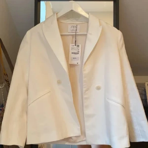 Vit kavaj från Zara - Snygg vit kavaj från Zara med klassisk krage och två knappar framtill. Jackan har två snedställda fickor och en rak, något croppad passform. Perfekt för en stilren look.