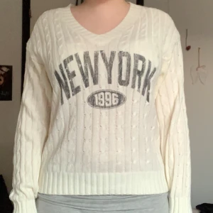 Vit stickad tröja med New York-tryck - Säljer en vit stickad tröja med kabelstickat mönster och stort grått 'NEW YORK 1996'-tryck framtill. Tröjan har en V-ringad hals och långa ärmar. Perfekt för en avslappnad och trendig look. Passar xxs-m! 