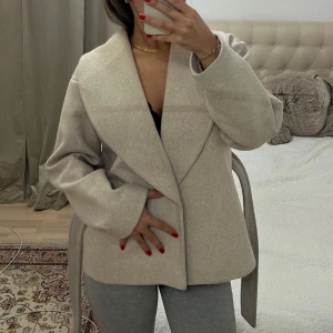 Beige kappa med bälte - Säljer en stilren beige kappa med bred krage och knytbälte i midjan. Jackan har en rak passform och är perfekt för dig som gillar en klassisk look. Passar till många olika outfits och har långa ärmar. Nypris 799, perfekt inför våren.