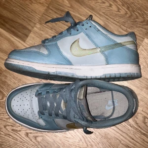 Nike Dunk sneakers i ljusblått och vitt - Säljer ett par Nike Dunk sneakers i ljusblått och vitt med guldfärgad swoosh. Skorna har klassisk snörning, perforerad tå och platt sula. Ganska använda därav priset. 