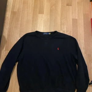 Svart tröja från Polo Ralph Lauren - Klassisk svart tröja från Polo Ralph Lauren med rund halsringning och den ikoniska röda loggan broderad på bröstet. Långärmad modell i mjukt material, perfekt för en stilren och enkel look.