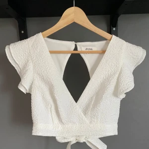 Vit croppad omlottopp med knyt - Säljer en vit croppad omlottopp från The Frolic med strukturmönster. Toppen har korta volangärmar, djup v-ringning både fram och bak samt knyt i midjan. 