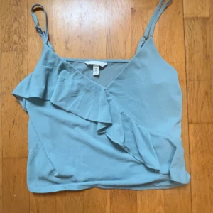 Ljusblå volanglinne från H&M - Säljer ett ljusblått linne från H&M med smala axelband och volangdetaljer framtill. Toppen har omlottkänsla och är perfekt för varma dagar. Enkel och stilren design som passar till mycket.