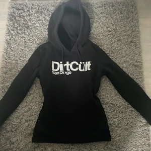 Svart hoodie från DirtCult  - Säljer en svart hoodie från DirtCult med vit logga på bröstet. Den är i storlek S men sitter mer som en XS. Den är nästan helt oanvänd och ser helt ny ut.