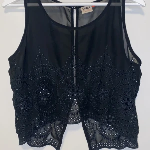 Svart topp från ONLY - Svart linnetopp från ONLY med broderade detaljer och hålmönster. Toppen är ärmlös och har knappar i ryggen.