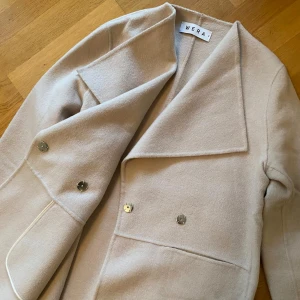 Ljusbeige ullkappa från Wera - Säljer en stilren ljusbeige kappa från Wera i ull. Jackan har guld färgade knappar så den kan stängas samt två större fickor. Perfekt för dig som gillar att hålla dig varm ute samt den klassiska looken.