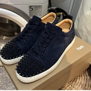 Mörkblå sneakers med nitar och röd sula - Säljer ett par mörkblå sneakers från Christian Louboutin med kromade nitar på tån och vit sula. Skorna har den ikoniska röda sulan och är tillverkade i mocka. Perfekta för dig som vill sticka ut med din stil. SNABB AFFÄR PRIORITERAS!!!