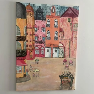 Målning av stadstorg med café och pastellfärgade byggnader - Säljer en färgglad 50x70 målning på duk som föreställer ett stadstorg med café, uteservering och pastellfärgade byggnader. Tavlan har detaljer som parasoller, blommor och smidesbalkonger. Perfekt för att skapa en mysig känsla i rummet.