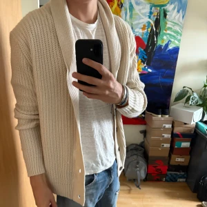 Beige stickad kofta  - Säljer en beige stickad kofta med sjalkrage och stora knappar framtill. Koftan har lång ärm och en grovstickad struktur som ger en avslappnad och stilren look. Perfekt att slänga på sig över en t-shirt.