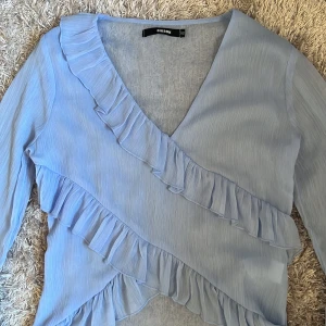 Ljusblå volangblus från Bik Bok - Supersöt ljusblå blus med omlott och volangdetaljer från Bik Bok. Blusen har långa ärmar med extra vida ärmslut och v-ringning. Perfekt för dig som gillar romantisk stil och vill sticka ut lite extra.