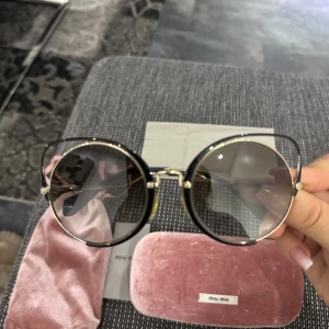 Runda solglasögon från Miu Miu - Snygga runda solglasögon från Miu Miu med tunn guldfärgad metallbåge och mörka glas. Kommer med rosa sammetsfodral och matchande tygpåse. Perfekt accessoar för att lyfta din stil.