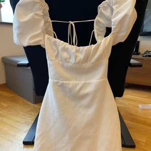 Vit klänning med puffärmar och knytband - Superfin vit klänning med puffiga kortärmar och snygga knytband både fram och bak. Klänningen har en feminin siluett och är perfekt för sommarens alla tillfällen. Materialet känns lätt och luftigt.