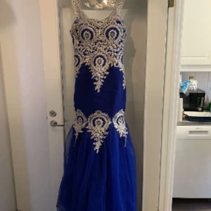 Blå långklänning med spetsdetaljer - Säljer en blå långklänning med vita spetsapplikationer och markerad midja. Klänningen har ärmlös design och figurnära passform upptill med utsvängd kjol i tyll. Perfekt för bal eller festliga tillfällen. En S/M som ny! Kan komma och provas också! 