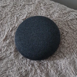 Google Home Mini Smarthögtalare - Säljer en Google Home Mini smarthögtalare i mörkgrått tyg med rund form. Högtalaren har tygöverdrag på ovansidan och plast på undersidan. Perfekt för att styra smarta hemmet, spela musik och få svar på frågor med röststyrning.