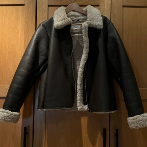 Brun pälsfodrad jacka med dragkedja - Säljer en brun jacka med mjukt beige foder och krage i fuskpäls. Jackan har dragkedja framtill och uppvikta ärmslut med samma pälsdetalj. Perfekt för kyliga dagar och har en klassisk, stilren look.