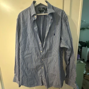 Blåvit randig skjorta från Polo Ralph Lauren - Säljer en blåvit randig skjorta från Polo Ralph Lauren.Skjortan har långa ärmar, klassisk krage och knäppning framtill. Broderad logga på bröstet. Perfekt för dig som gillar stilrena och klassiska plagg. Helt oanvänd och perfekt inför sommaren 