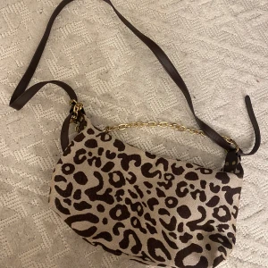 Leopardmönstrad axelväska med kedjedetalj - Säljer en snygg axelväska med leopardmönster i brunt och beige. Väskan har ett brunt axelband i syntet och en dekorativ guldfärgad kedja. Perfekt för att addera lite edge till din outfit.