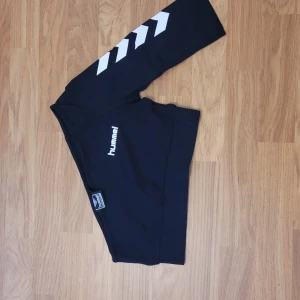 Svarta tights från Hummel med vita detaljer - Svarta tights från Hummel med vita pilar längs ena benet och Hummel-logga på låret. Perfekta för träning eller sportiga outfits. Stretchigt material som sitter tight och bekvämt.