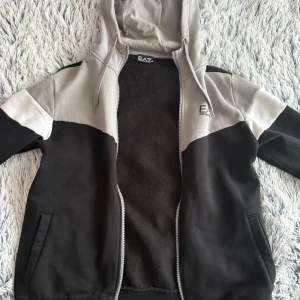 Svart och grå kofta från EA7 Emporio Armani - Säljer en svart och grå hoodiejacka från EA7 Emporio Armani. Jackan har dragkedja, huva och två fickor framtill. Perfekt för en sportig och avslappnad stil.
