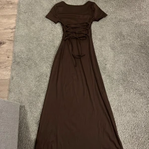 Brun långklänning med snörning i midjan - Säljer en brun långklänning med korta ärmar och rund halsringning. Klänningen har en snygg snörningsdetalj i midjan som ger en figurnära passform. Perfekt för dig som gillar en enkel men stilren look.