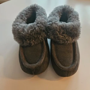 Gråa mysiga tofflor med fuskpäls - Supergosiga gråa tofflor med fluffig fuskpäls runt öppningen. Perfekta att ha hemma för att hålla fötterna varma under kyliga dagar. Mjukt material och klassisk design. Storlek 35. Knappt använda.