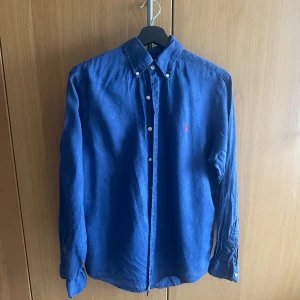 Blå linneskjorta från Ralph Lauren - Säljer en klassisk blå skjorta i linne från Ralph Lauren med vit knappstängning och röd broderad logga på bröstet. Skjortan har lång ärm och button-down krage. Perfekt för en stilren och avslappnad look. Står storlek S men skulle säga att den är väldigt loose fit så passar bra för någon som normalt har Medium.