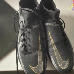 Svarta Nike fotbollsskor med gula detaljer/NIKE PHANTOM GT 2 ELITE FG SHADOW - SVART/GRÅ/GULD - Säljer ett par svarta Nike fotbollsskor med stora gula swoosh-loggor och snörning. Skorna har rund tå och platt sula med dobbar, perfekta för spel på gräsplan. Snygg design med silverdetaljer längs sidorna. Inga skador utöver linjerna på bilden.