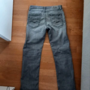 Grå jeansbyxor från Replay - Säljer ett par grå jeansbyxor från Replay med klassisk femficksdesign och raka ben. Byxorna har diskreta slitningar och tydliga sömmar på bakfickorna. Perfekta för dig som gillar en stilren och avslappnad look.