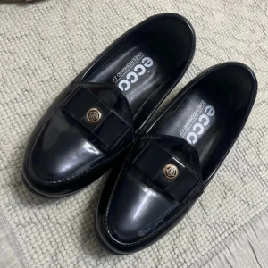 Svarta loafers från Ecco med dekorativ detalj - Stilrena svarta loafers från Ecco med glansig finish och dekorativ rosett samt guldfärgad knapp framtill. Skorna har rund tå och platt sula, perfekta för en klassisk look. Passar dig som gillar tidlös design och komfort.
